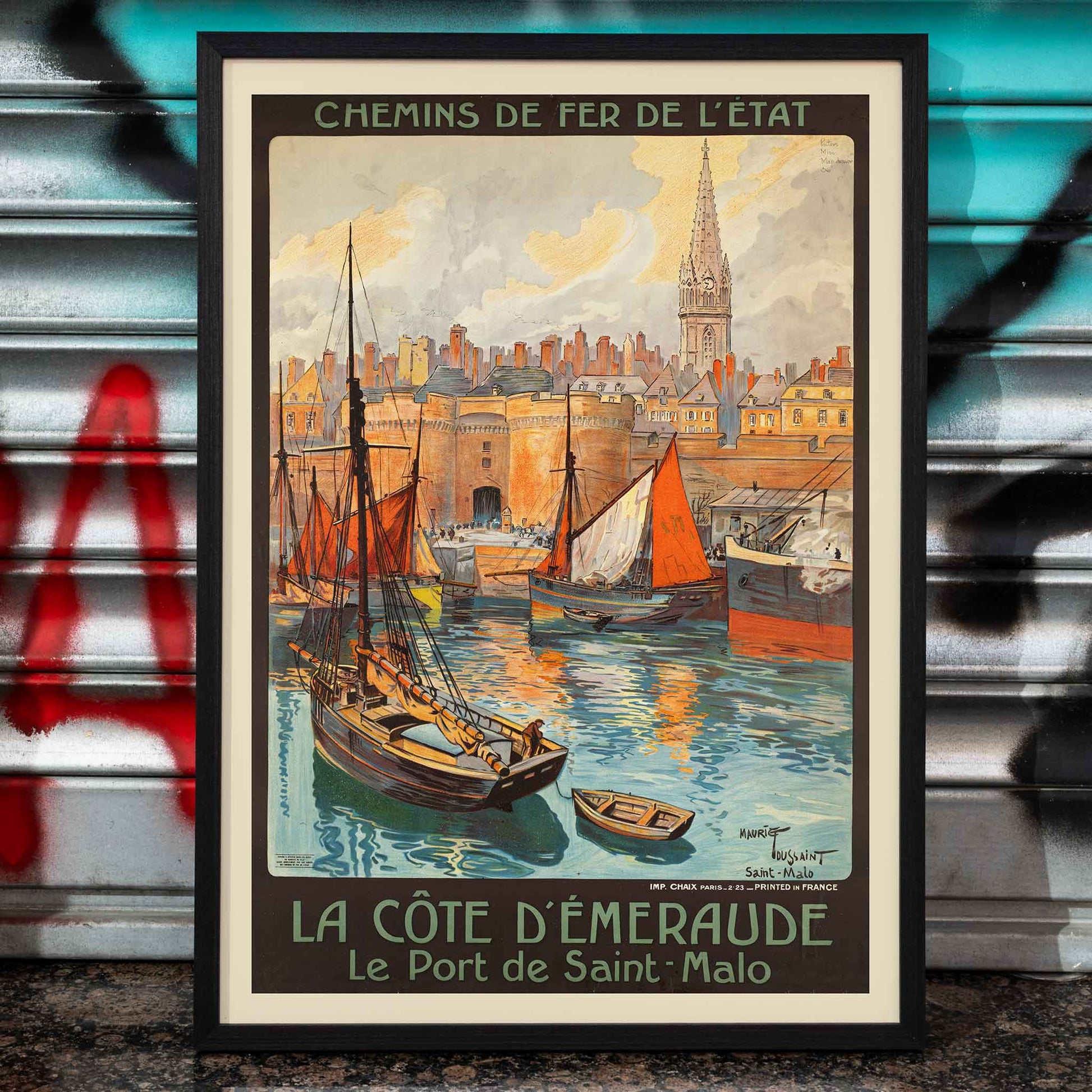 Colorful vintage travel poster of Saint-Malo harbor titled La cote d’Emeraude Le Port de Saint-Malo by Mourgue and Maurice Toussaint featuring sailing boats in a vibrant harbor scene, framed and displayed in home decor