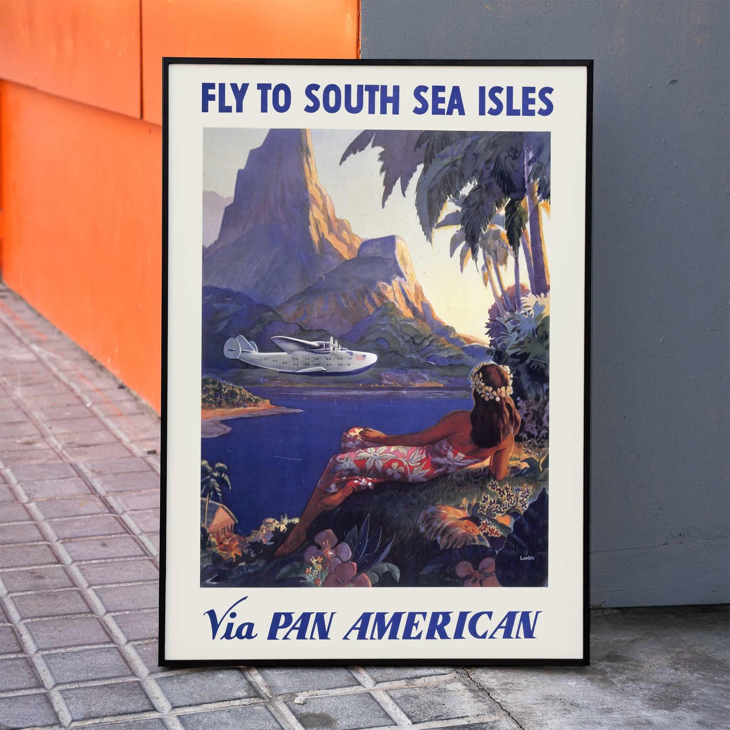 Vintage travel poster of a woman in a floral dress on a tropical island with a Pan American seaplane by Paul George Lawler, framed and displayed in home decor