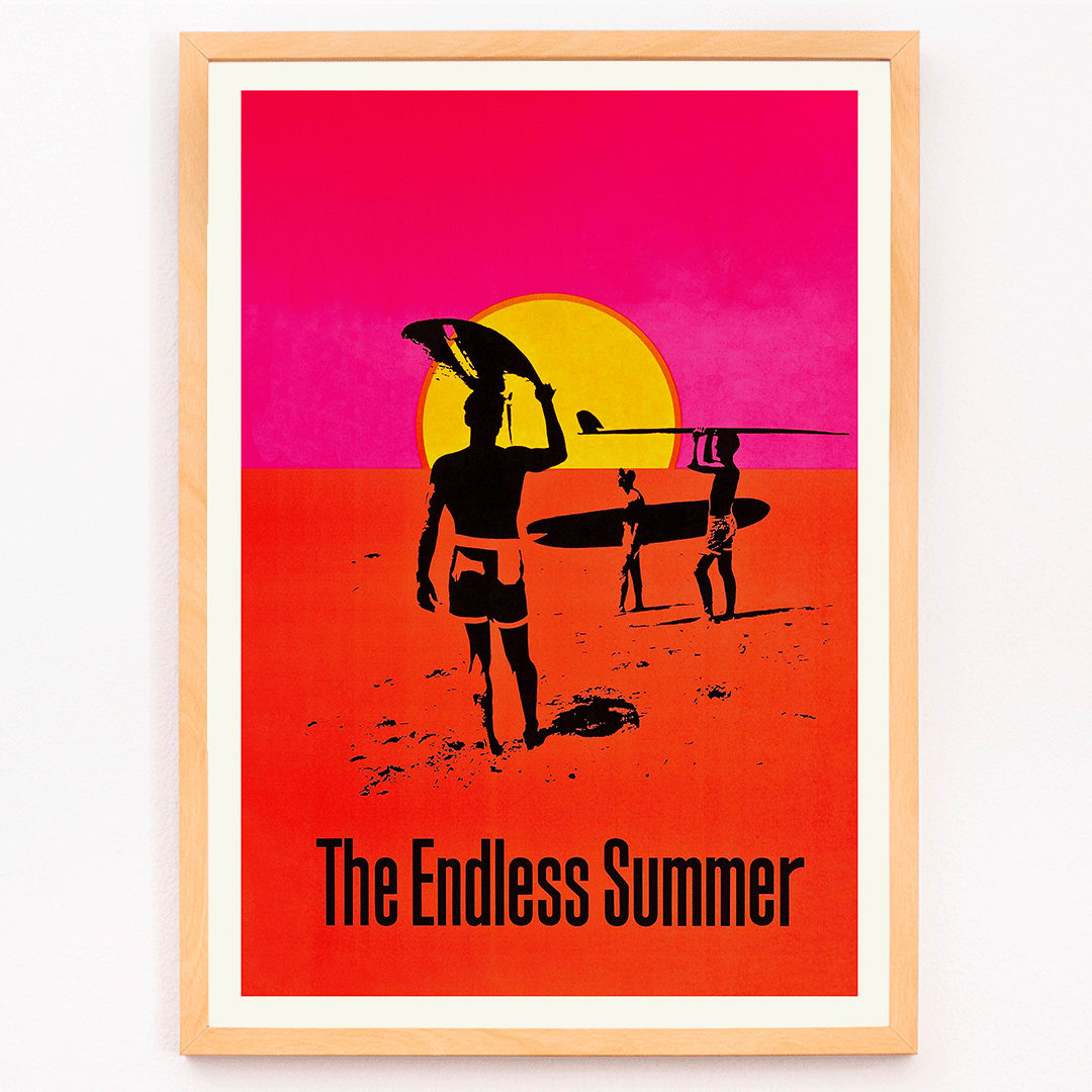 The Endless Summer poster with surfers carrying boards on a beach at sunset against a stylized sunset backdrop, framed and displayed in modern living room decor